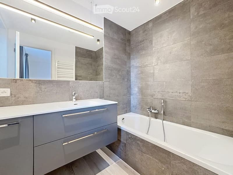 Modern bathroom with grey tiled walls, a white bathtub, a sink with a modern faucet, and a large mirror with built-in lighting.