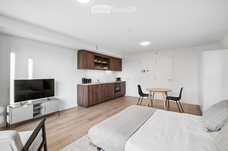Modern, open-plan living and sleeping area with a bed, small dining table with chairs, and a fitted kitchen with cabinets, sink, oven, and fridge. The space features light wooden flooring, white walls, and recessed lighting.