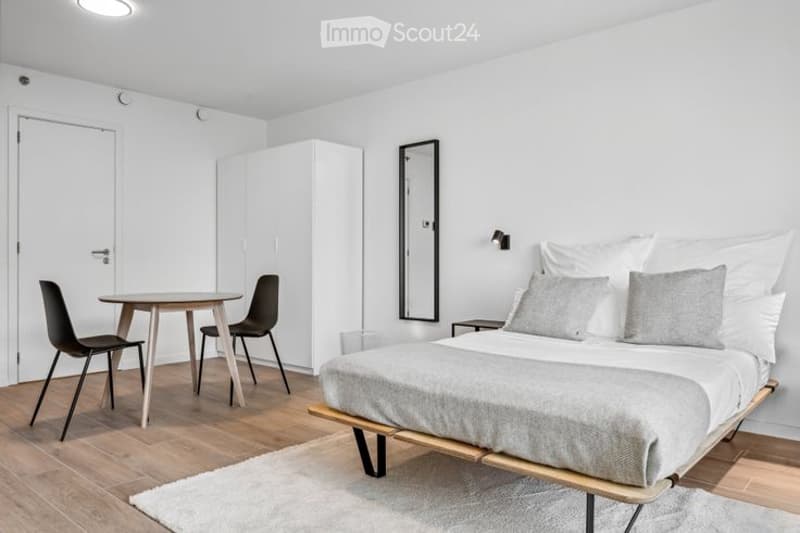 Modern, minimalist bedroom with a bed, small round table, chairs, a mirror, and a wardrobe. The room features light wooden flooring and white walls.