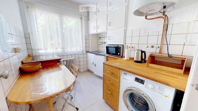 A compact kitchen with white cabinets, wooden countertops, a small dining table with a chair, a microwave, a kettle, and a washing machine.