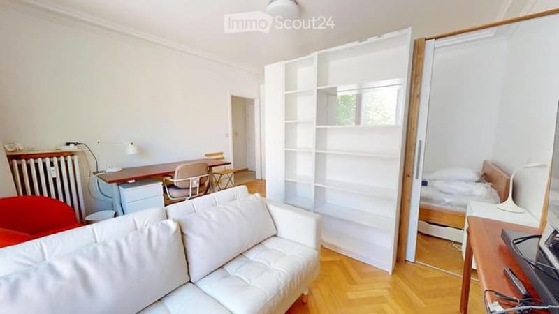 A modern, well-lit living space with a white sofa, a wooden floor, a bookshelf, a desk, and a bed in the background.