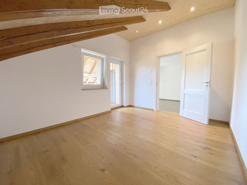 Empty room with light wooden flooring, white walls, exposed wooden beams on the ceiling, and a window with a view of another room.