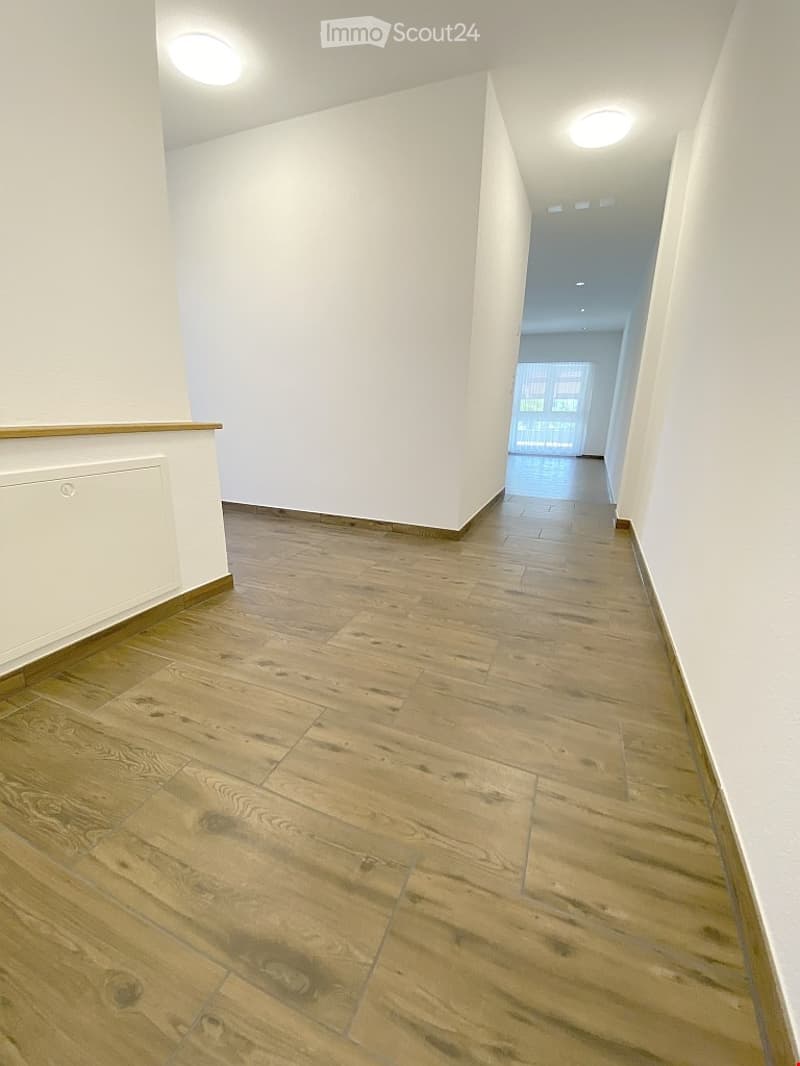 Empty hallway with light wooden flooring, white walls, and recessed ceiling lights. The hallway leads to an open area with large windows and a view of greenery.