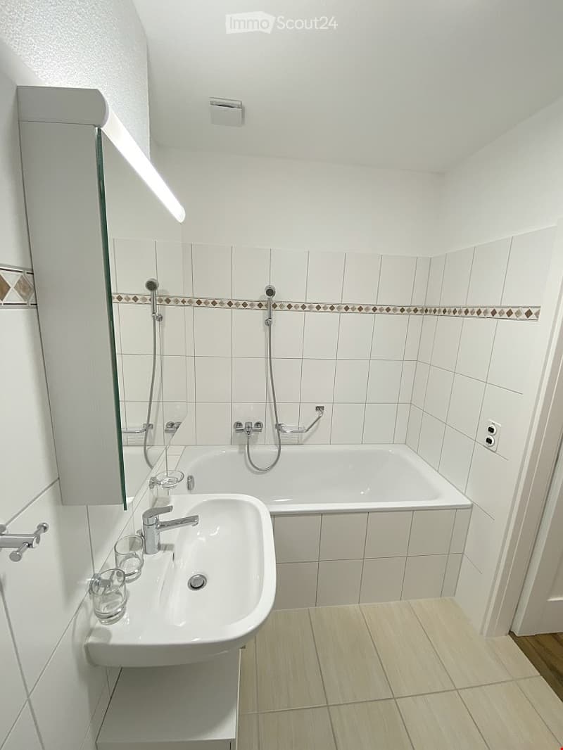 Modern bathroom with white tiles, a sink, bathtub, and a large mirror with built-in lighting.
