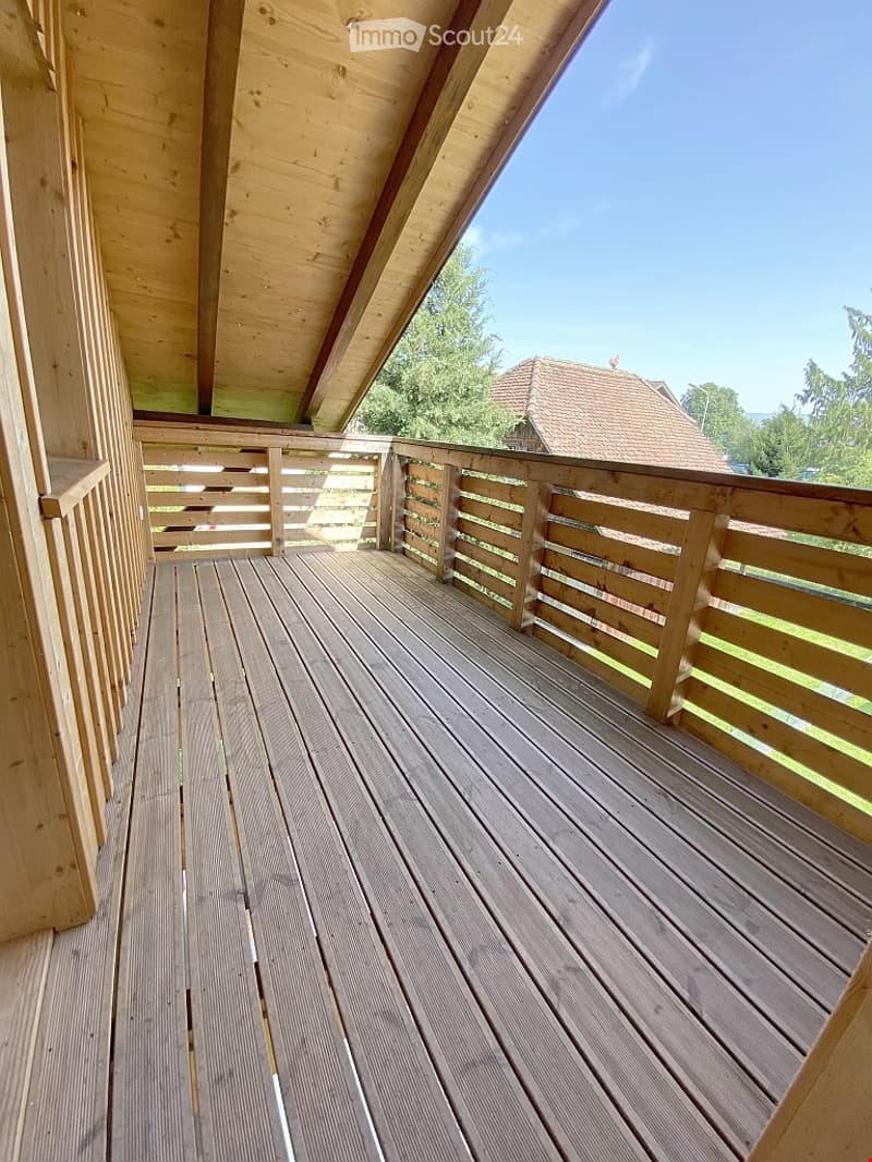 Wooden balcony with slatted flooring and wooden railings, featuring a clear blue sky and distant house in the background.