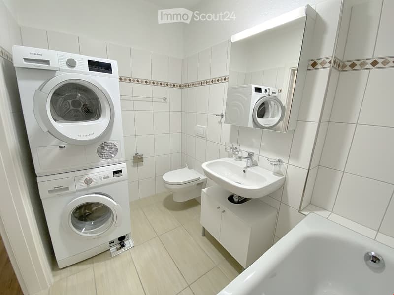 Modern bathroom with white tiles, a toilet, sink, bathtub, and stacked washing and drying machines.