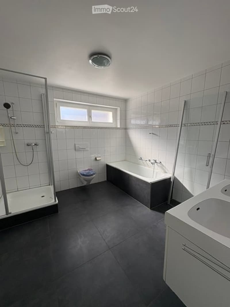 Modern bathroom with white tiled walls, a bathtub, toilet, sink, and a glass-enclosed shower stall. The room features a large window for natural light and a ceiling light fixture.