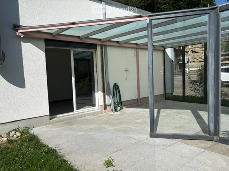 A modern glass-covered patio area attached to a house, with a sliding glass door, tiled floor, and a metal awning.