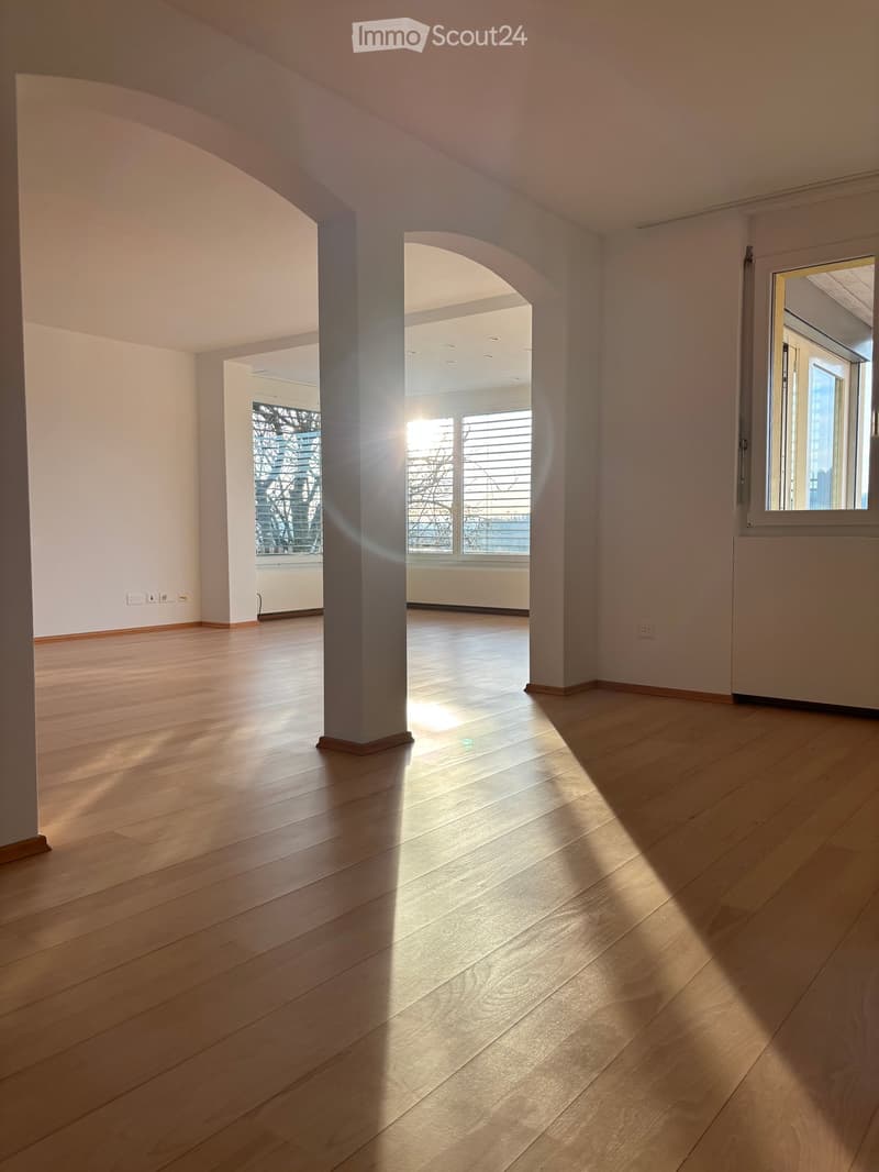 Empty room with wooden flooring, white walls, and large windows allowing natural light to enter. The space is open and bright with visible architectural columns and electrical outlets.