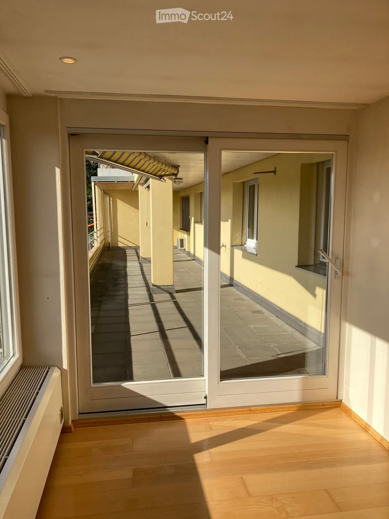 Bright hallway with wooden flooring, large glass doors leading to a sunlit outdoor area, white walls, and a radiator on the left side.