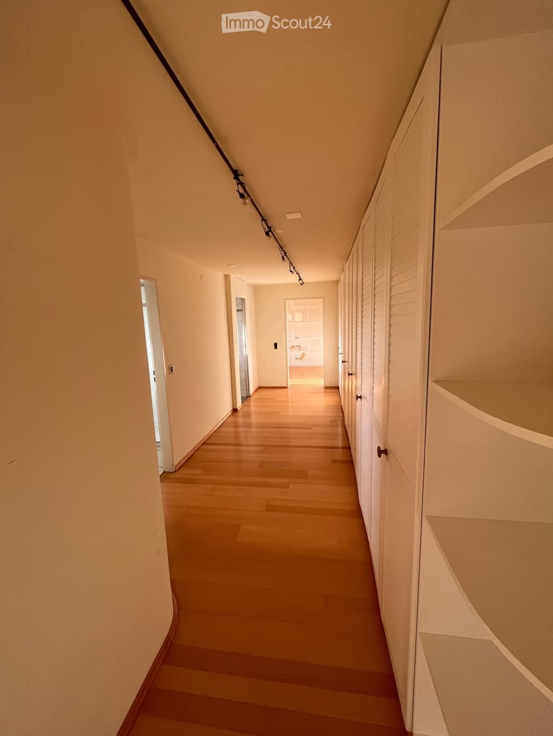 A long, narrow hallway with light wooden flooring and white walls, featuring several closed doors and recessed lighting fixtures.