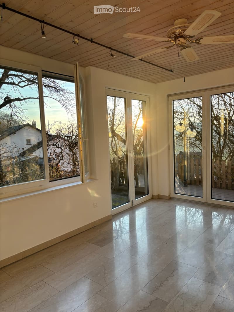 Bright, empty room with large windows and sliding glass doors leading to a wooden deck, featuring a polished marble floor and a wooden ceiling with recessed lighting.