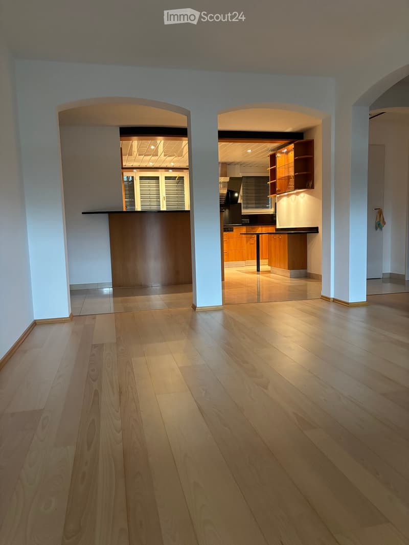Empty room with light wooden flooring, white walls, and two open archways leading to a kitchen area with wooden cabinets and a countertop.