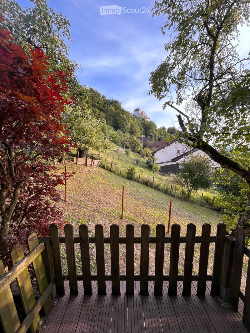 Wooden deck with a wooden fence overlooking a hillside with trees, a house, and a distant castle-like structure.