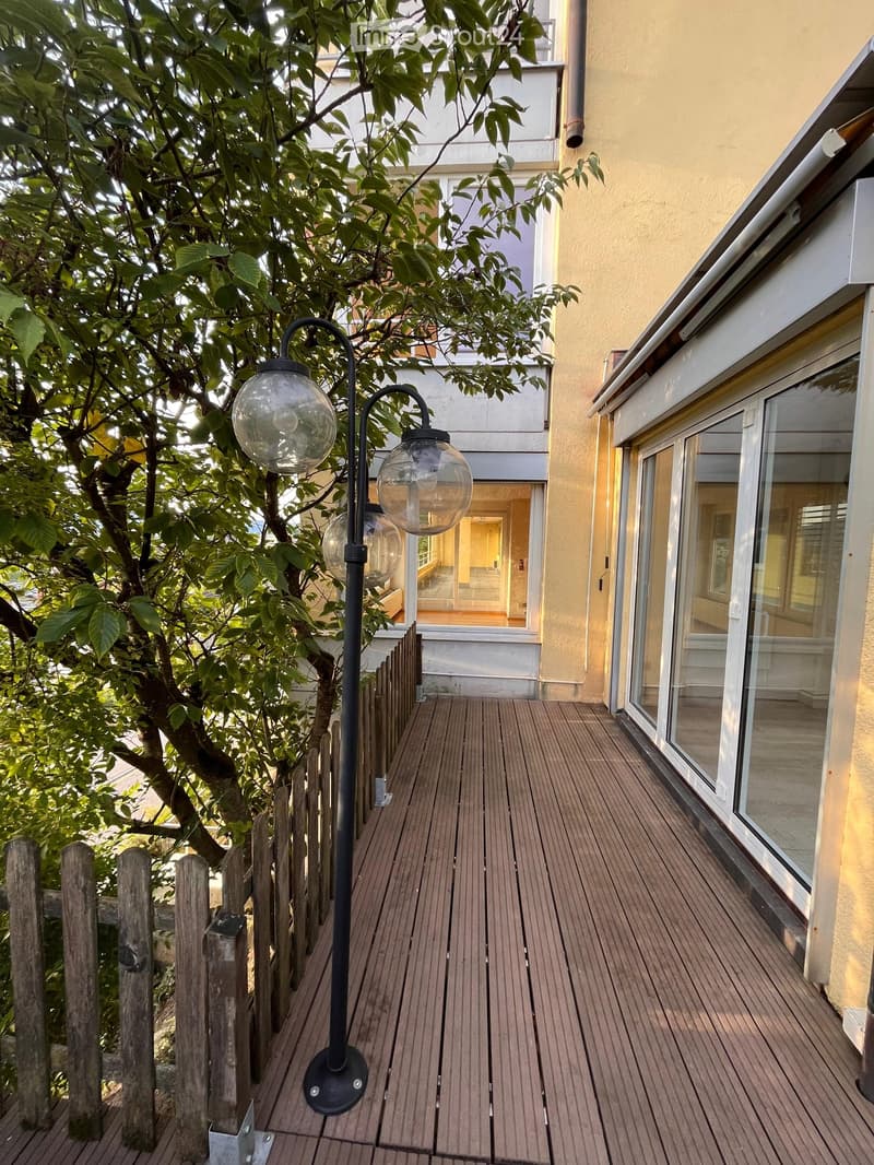 Wooden deck with a black lamp post featuring glass globes, adjacent to a building with large windows and a small wooden fence.