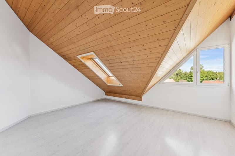 Bright, empty room with white walls, wooden ceiling, and large windows offering a view of greenery. The floor is covered with light-colored tiles.