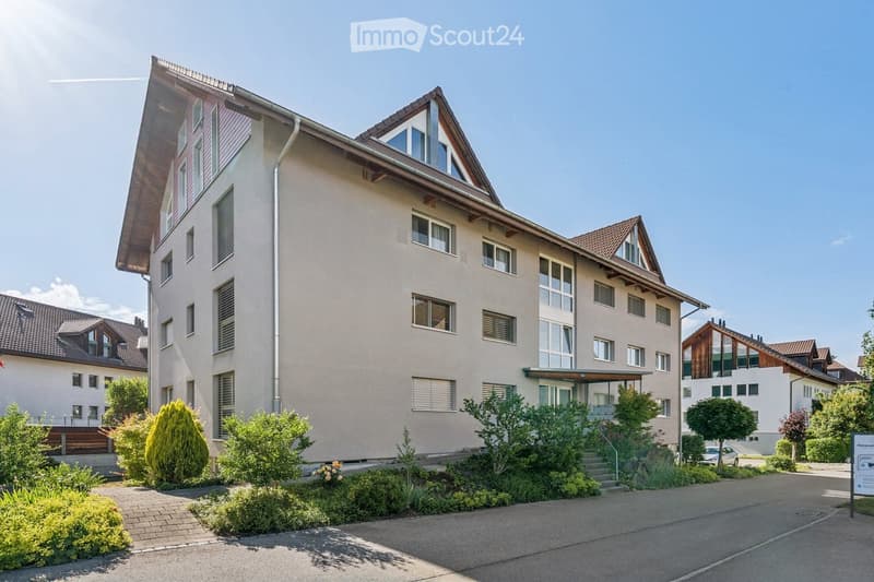 Modern multi-story residential building with light-colored facade, multiple windows, and a well-maintained garden area in front.
