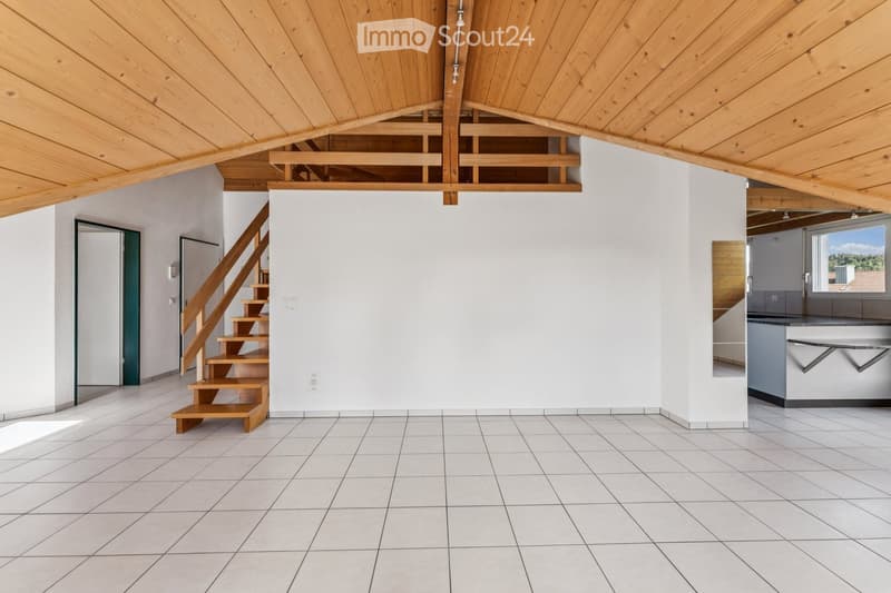 Empty room with white tiled floor, white walls, wooden staircase leading to an upper level, and a view of a kitchen area with modern cabinets and a window.