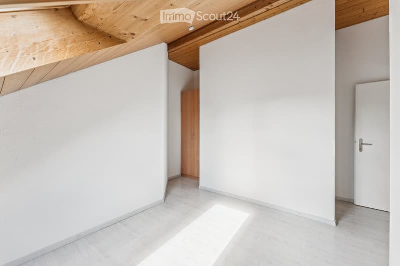 Empty room with white walls, light wooden ceiling, and light-colored floor. Features a small wooden cabinet and a white door.