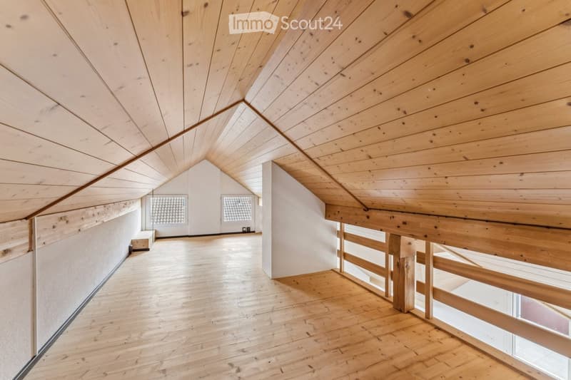 An empty attic space with wooden floors and exposed wooden beams, featuring a wooden railing and two windows with frosted glass panels.