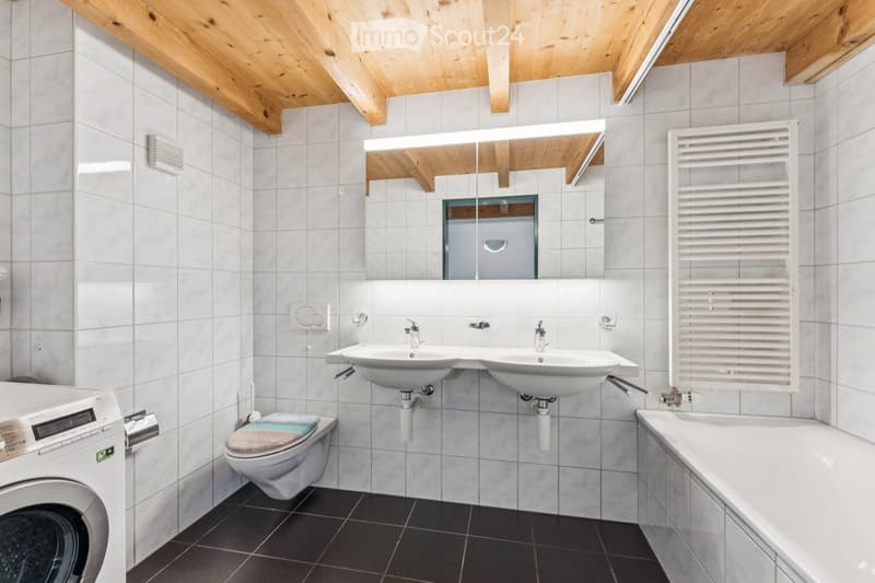 Modern bathroom with white tiles, double sink, toilet, bathtub, radiator, and washing machine. Features a wooden slanted ceiling and a large mirror above the sink.