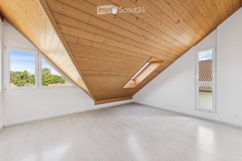 Bright, empty attic room with wooden slatted ceiling, white walls, light wooden floor, and large windows offering natural light and views of the outdoors.