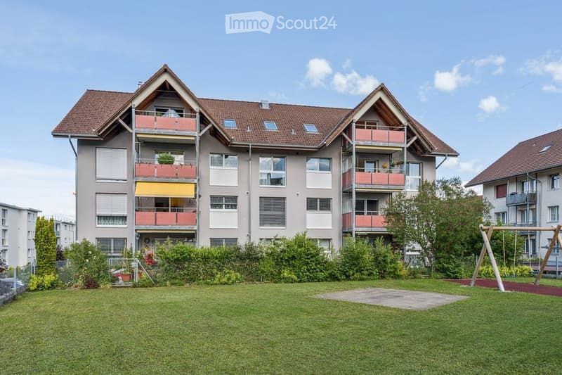 Multi-story residential building with balconies, surrounded by a well-maintained lawn and playground equipment.