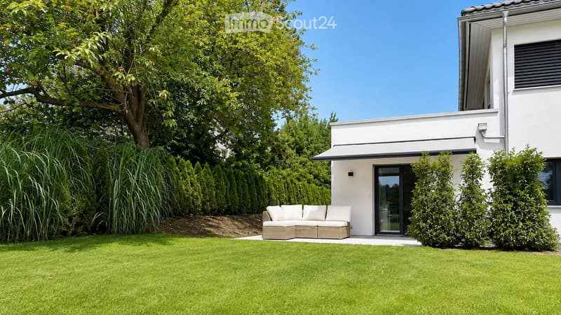 Modern outdoor seating area with a beige sofa and white cushions, positioned on a well-maintained lawn in front of a white house with large windows and a covered entrance.