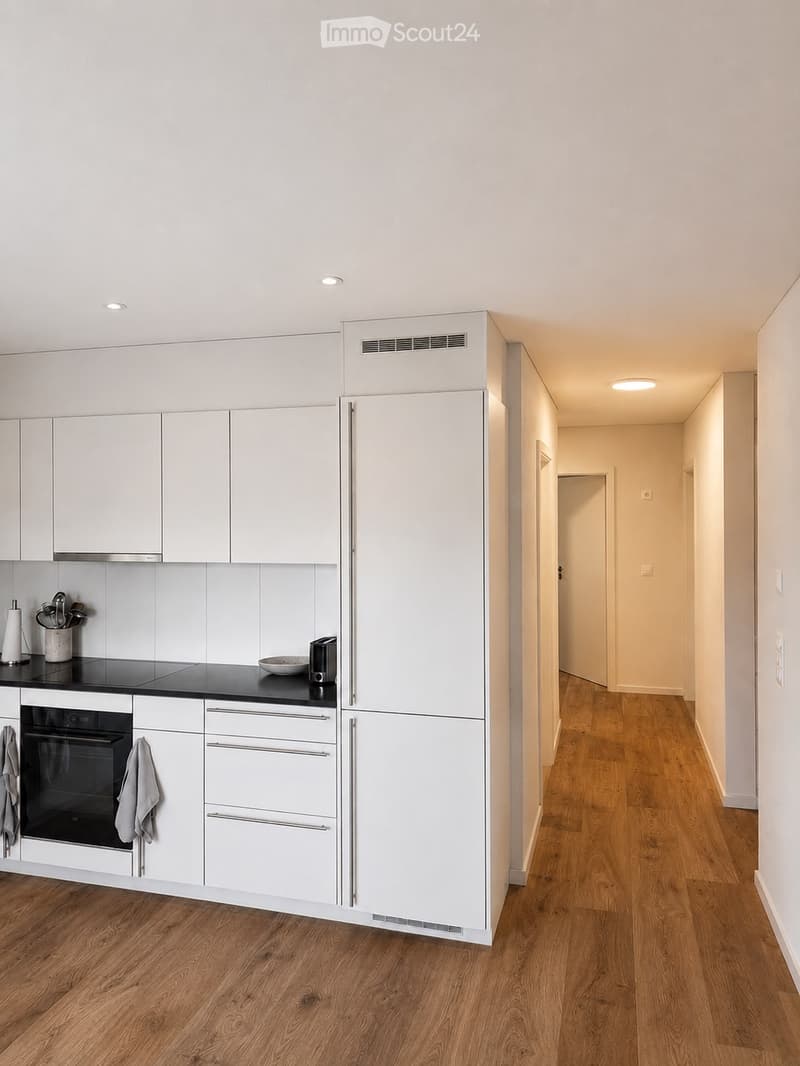 Modern hallway with wooden flooring, white walls, and recessed lighting. The hallway leads to a kitchen area with white cabinets, stainless steel appliances, and a black countertop.
