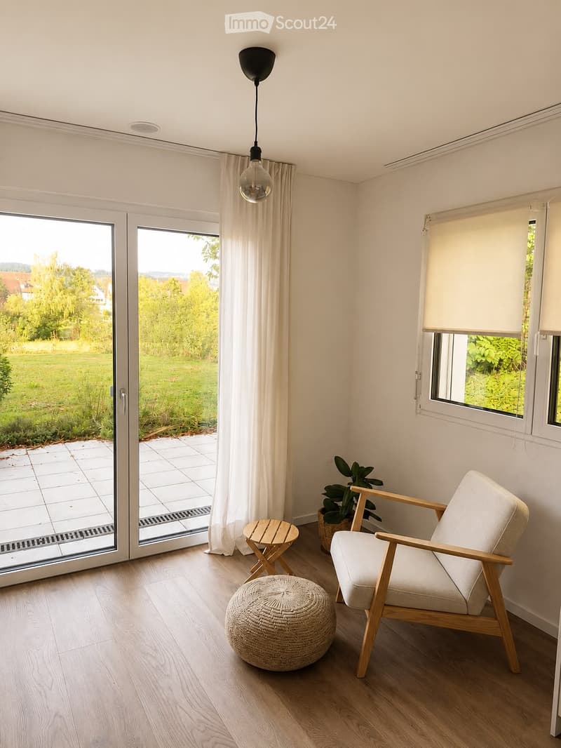 Bright living room with large sliding glass doors leading to a patio, wooden floor, white walls, a hanging light fixture, a round ottoman, a wooden chair, a small wooden table, and a potted plant.