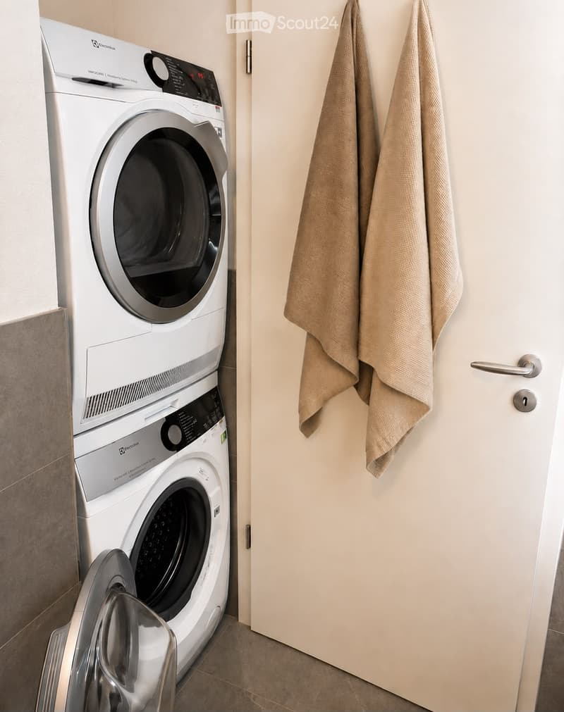 Modern bathroom with a stacked washing machine and dryer unit, beige towels hanging on the wall, and a white door.