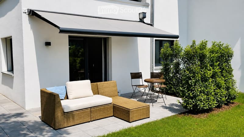 Outdoor patio with a wicker sofa, white cushions, a small round table, and chairs, adjacent to a modern house with a retractable awning and large sliding glass doors.