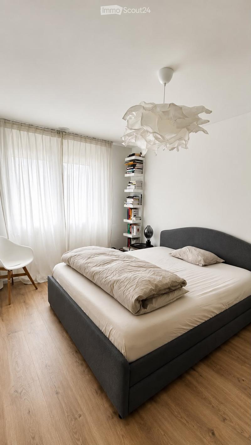 Modern bedroom with a large bed, white bedding, a wooden floor, a white chair, a bookshelf, and a decorative lamp.