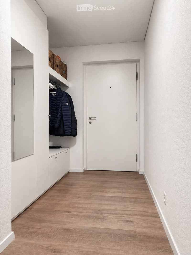 A narrow hallway with light wooden flooring, white walls, and a built-in closet on the left side. The closet contains hanging jackets and a small shelf with baskets. A closed white door is at the end of the hallway.