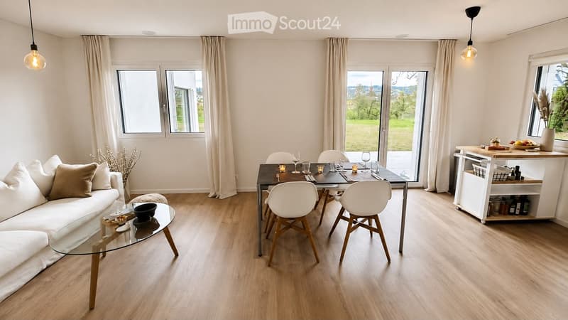 Modern living and dining area with light wood flooring, large windows, a white sofa, glass coffee table, dining table with chairs, and a bar cart.