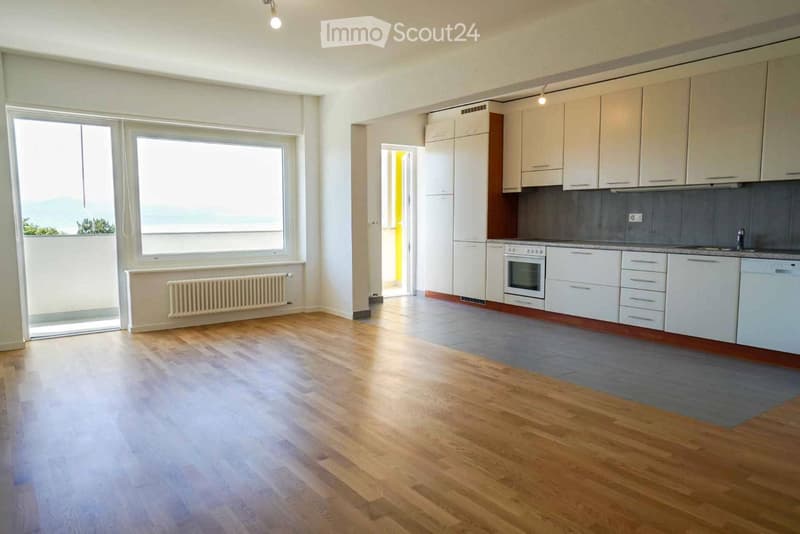 Empty room with wooden flooring, large windows, radiator, and an open doorway leading to a kitchen area with white cabinets and appliances.