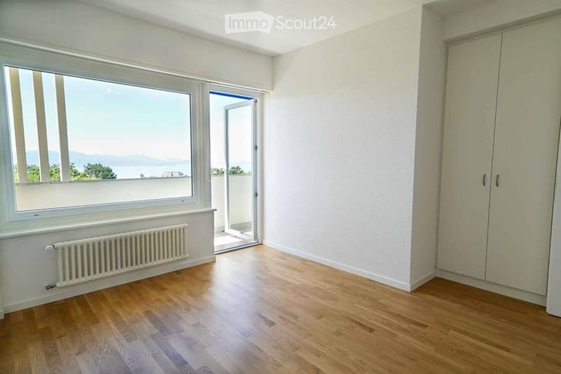 Empty room with wooden flooring, white walls, large window with scenic view, built-in wardrobe, and radiator under the window.