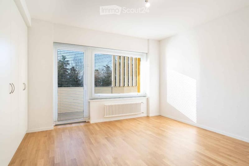 Empty room with light wooden flooring, white walls, large windows with blinds, and a radiator below one window.