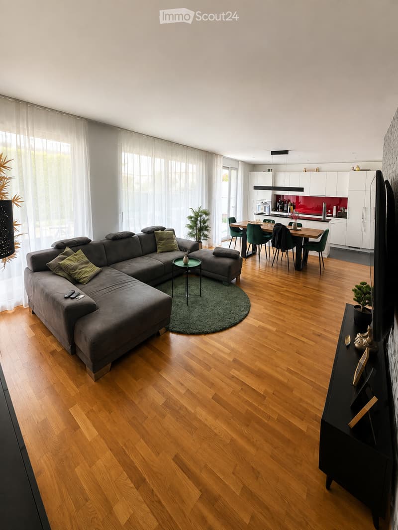 Open-plan living and dining area with hardwood flooring, large windows with white curtains, grey sectional sofa, coffee table, dining table with chairs, and a modern kitchen with white cabinets and red backsplash.