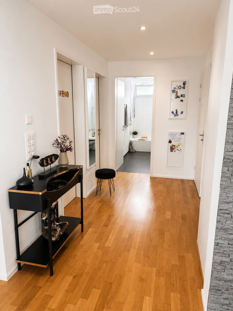 Modern hallway with wooden flooring, white walls, and recessed lighting. Features a black console table with decorative items, a small black stool, and a view into a bathroom with a bathtub and white tiles.
