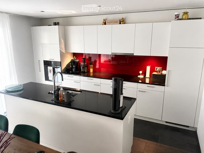 Modern kitchen with white cabinets, black countertops, and a red backsplash. Features a sink, faucet, oven, coffee maker, blender, knife block, and various small appliances.