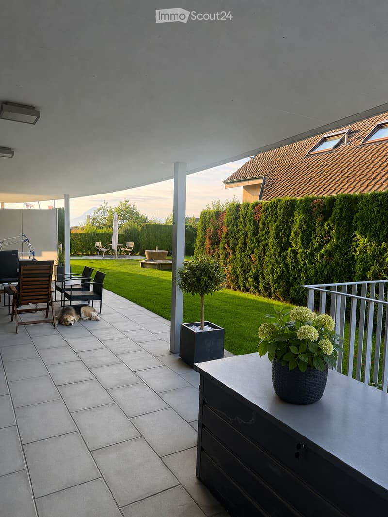A modern outdoor patio with tiled flooring, a small table with a vase of flowers, a dog resting, and a well-maintained lawn with surrounding greenery and a hedge.