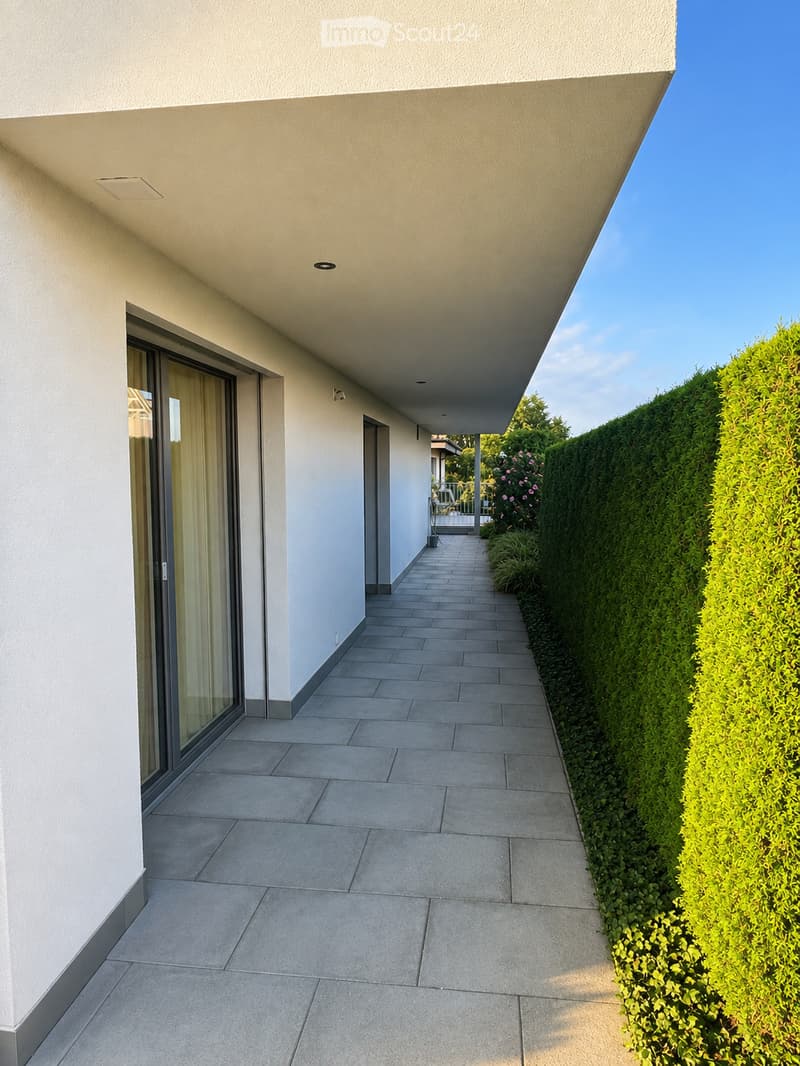 A modern covered walkway with tiled flooring, white walls, and a large glass door on one side. The walkway is bordered by neatly trimmed green hedges and leads to an outdoor area.