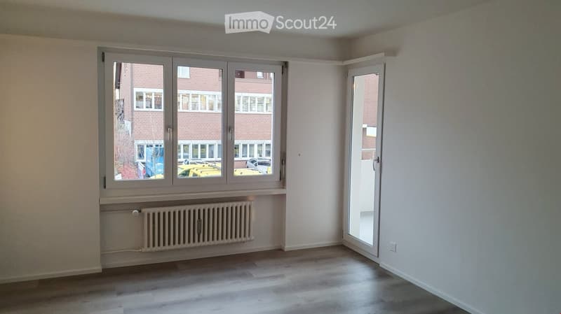 Empty room with light-colored walls, wooden flooring, large window with radiator below, and an open door leading to a balcony or outdoor area.
