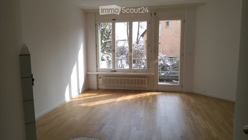 Empty room with wooden flooring, large windows, and a sliding glass door leading to a balcony. The walls are painted white, and there is a radiator below the windows.