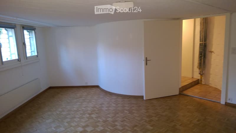 Empty room with wooden flooring, white walls, and two windows. A door leads to another area with tiled flooring.