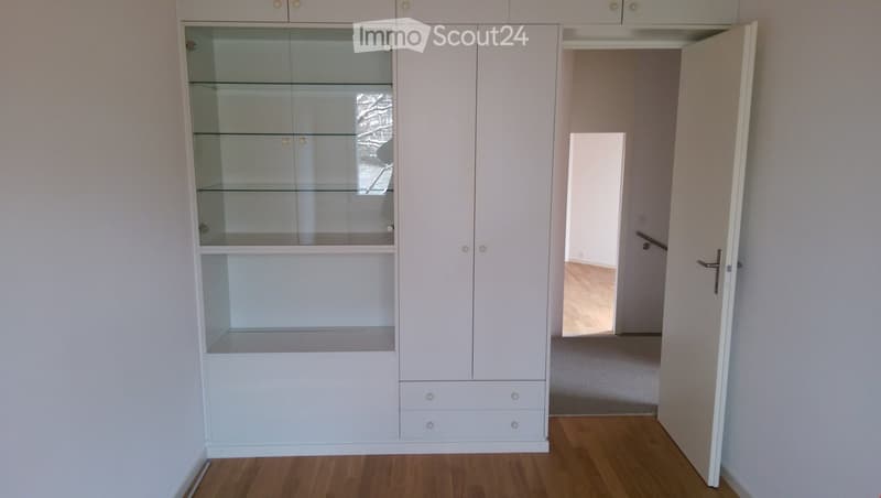 Empty room with light wooden flooring, white walls, and a large white cabinet with glass doors and drawers. An open doorway leads to another room with similar flooring.