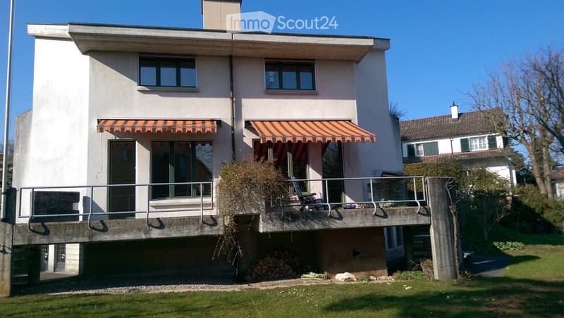 Two-story house with white exterior, large windows, balconies with striped awnings, and a small garden area.