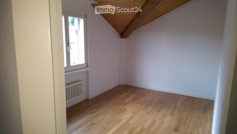 Empty room with wooden flooring, white walls, and a slanted wooden ceiling. A window with a radiator below is visible on one wall.