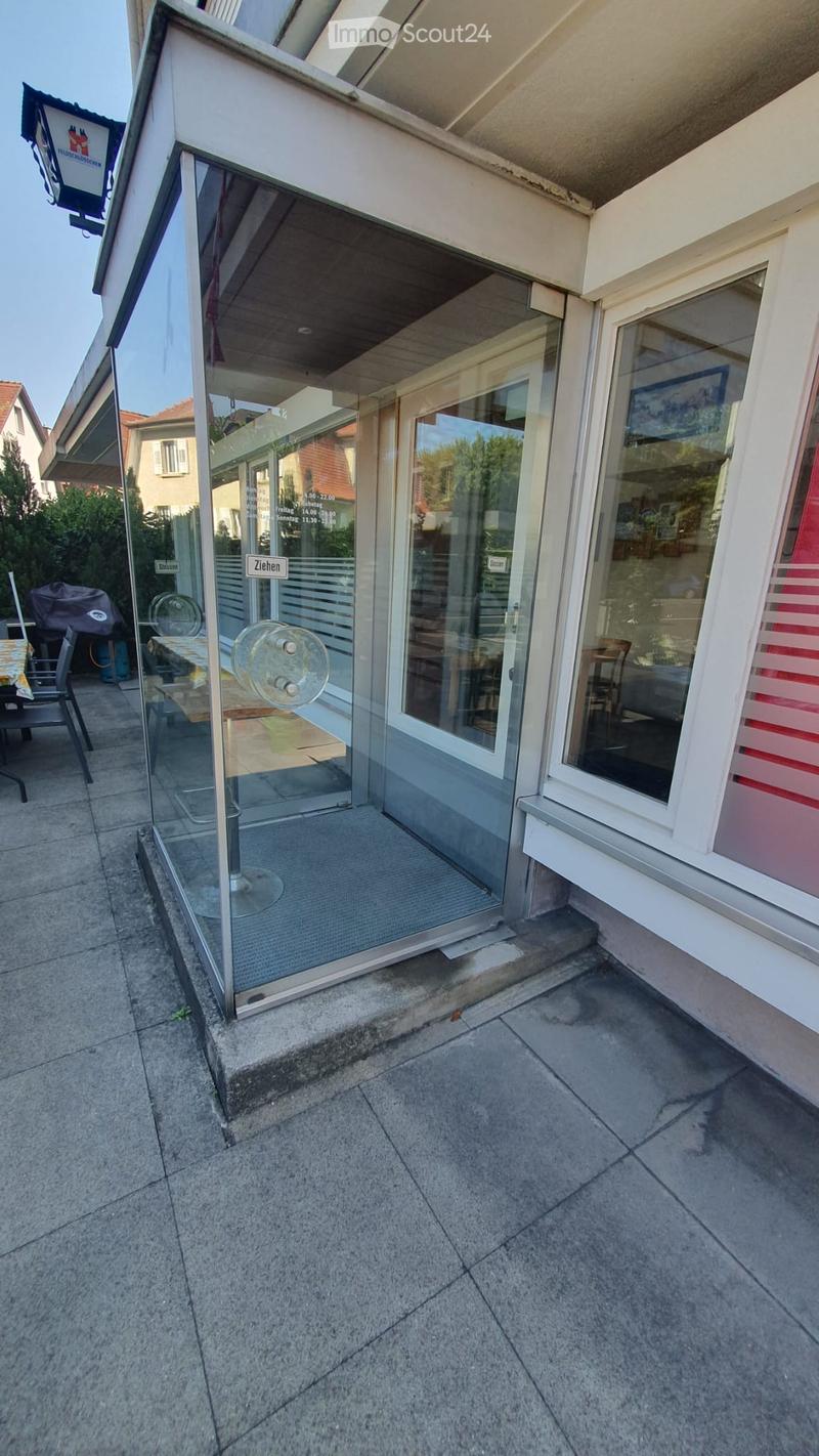 Outdoor patio area with a glass-enclosed entrance to a building, featuring a table, chairs, and a fan. The area is paved with concrete tiles and includes a sign indicating "Ziehen" (moving).